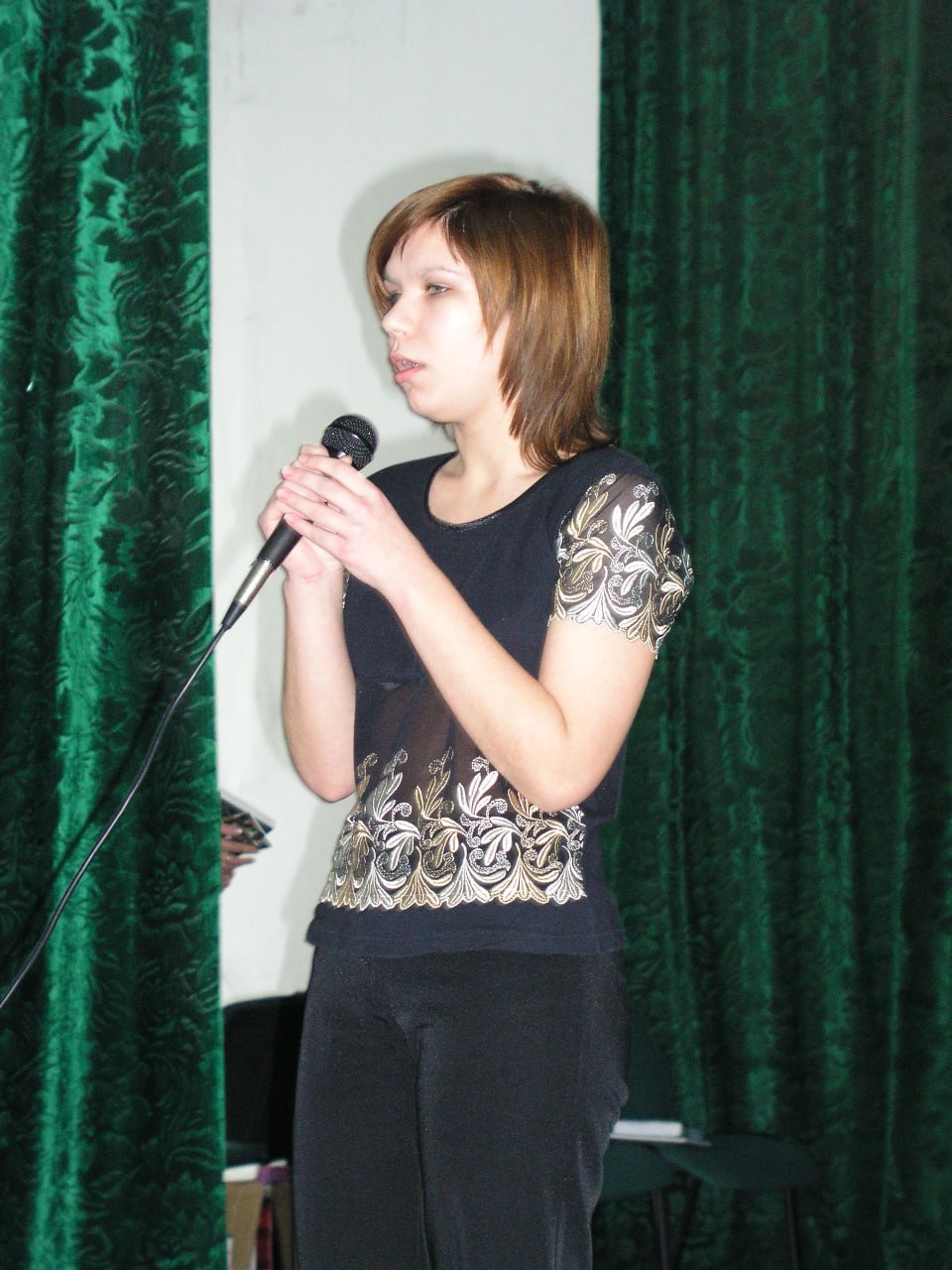 Женщина выступает на сцене с микрофоном, одета в черное платье с вышивкой, фон с зелеными шторами.